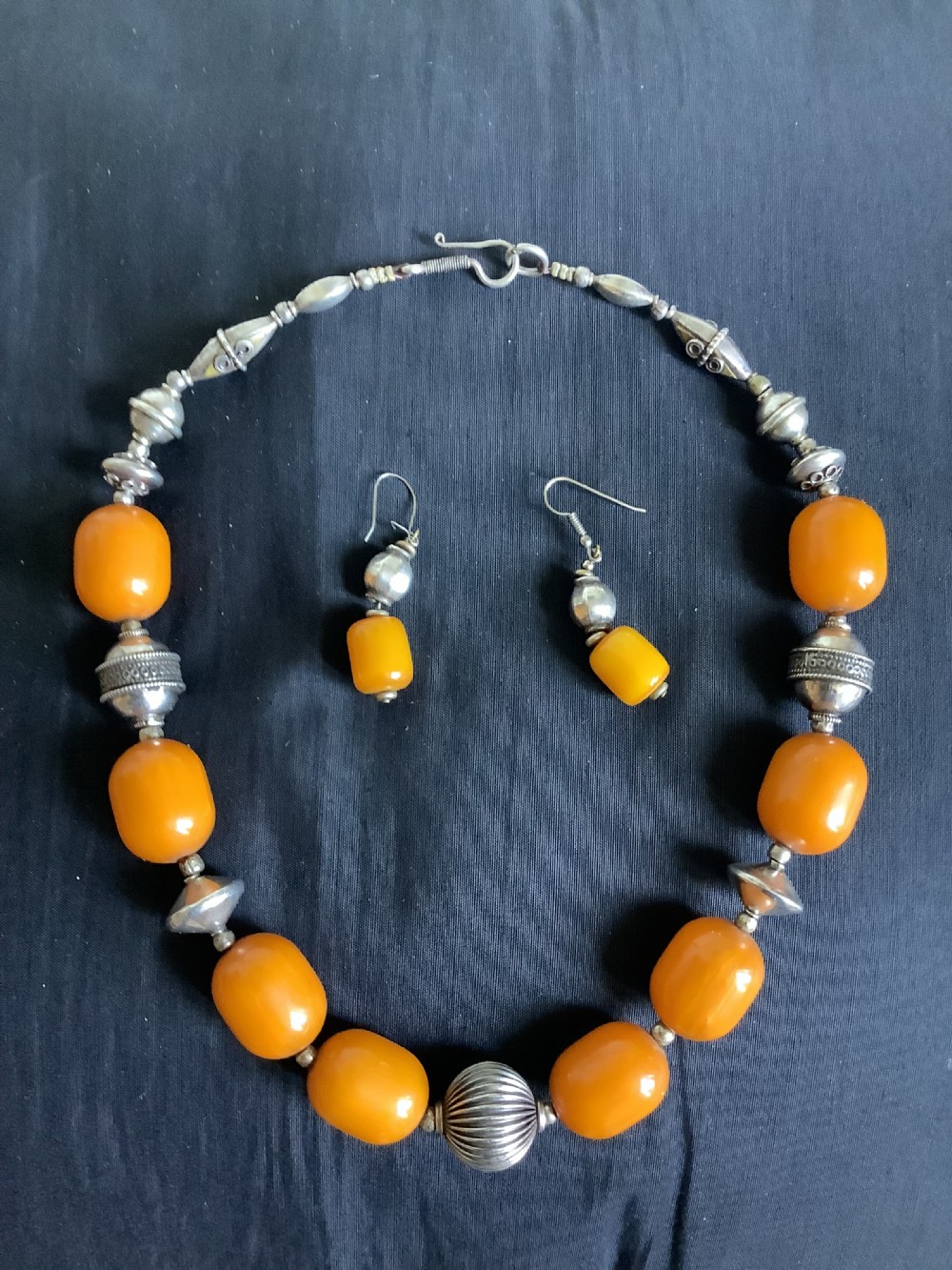 An orange amber bead and sterling silver necklace with matching drop earrings displayed flat on a dark fabric surface. View VI