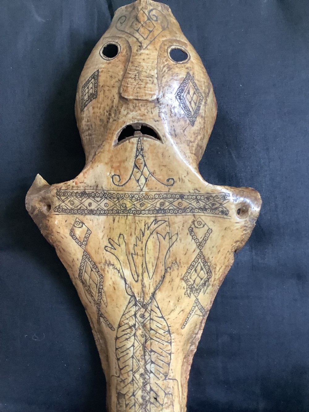 A carved beige animal bone featuring stylised human facial details, incised geometric diamond patterns, and an engraved reptile design. View V