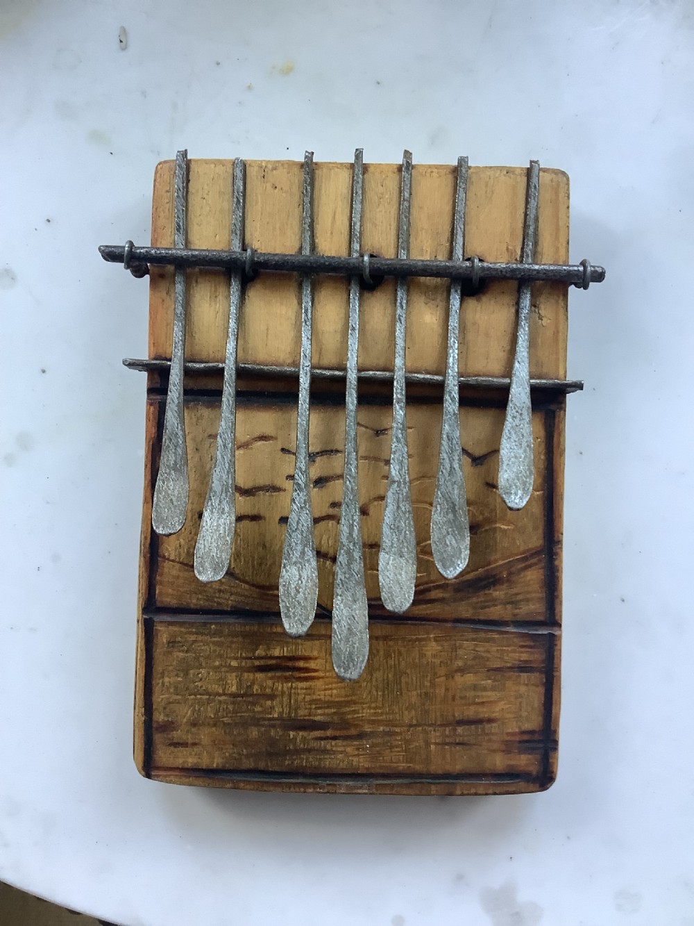 A rectangular wooden African mbira thumb piano with seven hand-forged metal tines set over a burnt decorated wood soundboard.