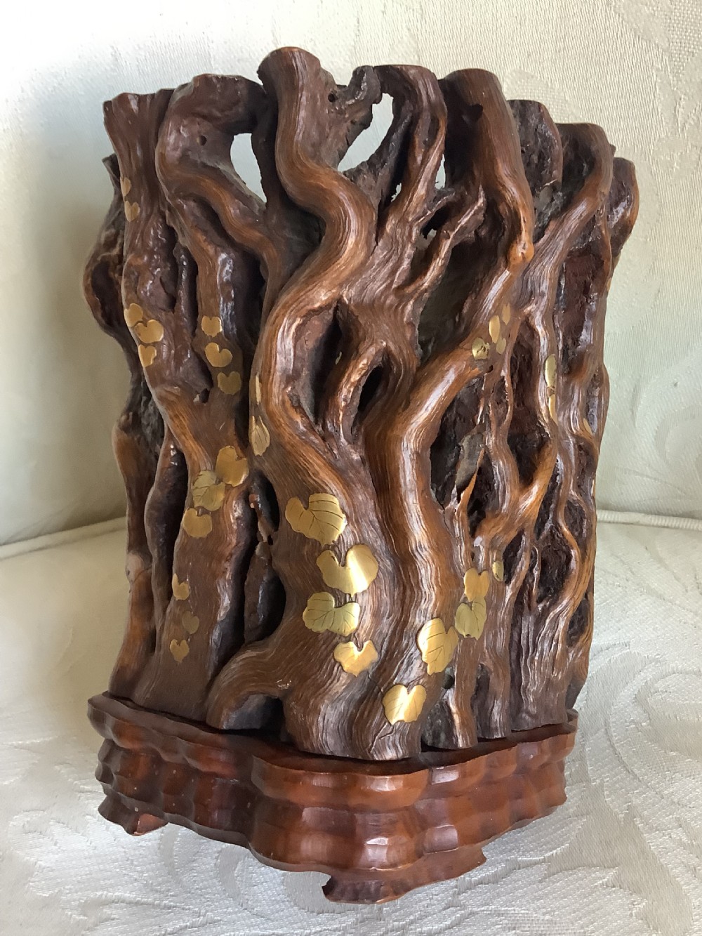 A cylindrical Chinese carved hardwood brush pot featuring openwork gnarled vine motifs inlaid with brass leaves, resting on a fitted carved wooden base. View X