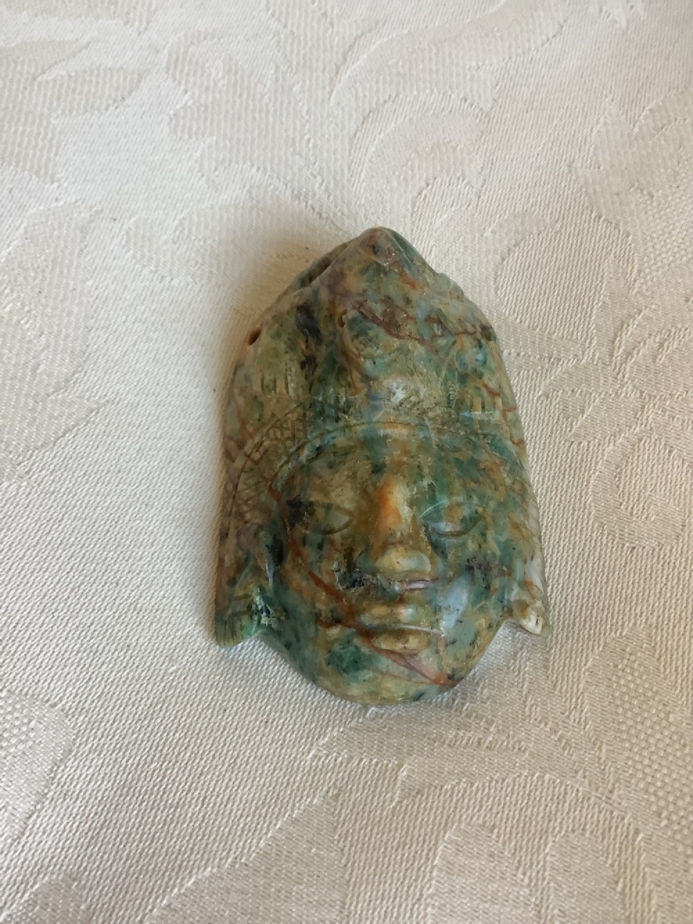A carved mottled green and brown jade pendant depicting a stylized human face with an ornate carved headdress.