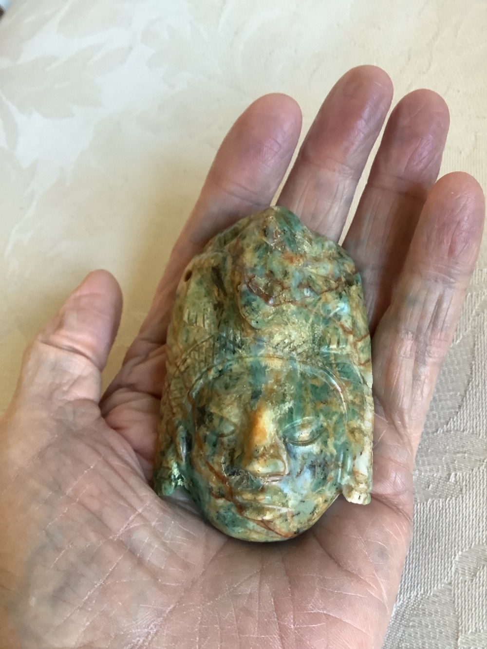 A carved mottled green and brown jade pendant depicting a stylized human face with an ornate carved headdress. View II