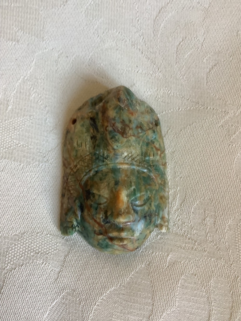 A carved mottled green and brown jade pendant depicting a stylized human face with an ornate carved headdress. View III