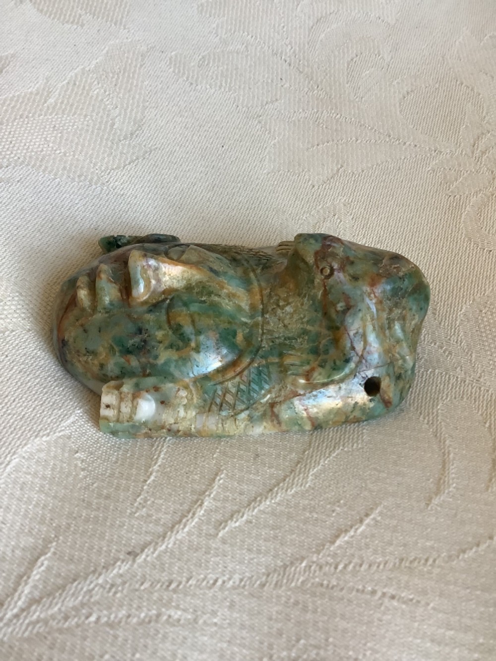 A carved mottled green and brown jade pendant depicting a stylized human face with an ornate carved headdress. View IV