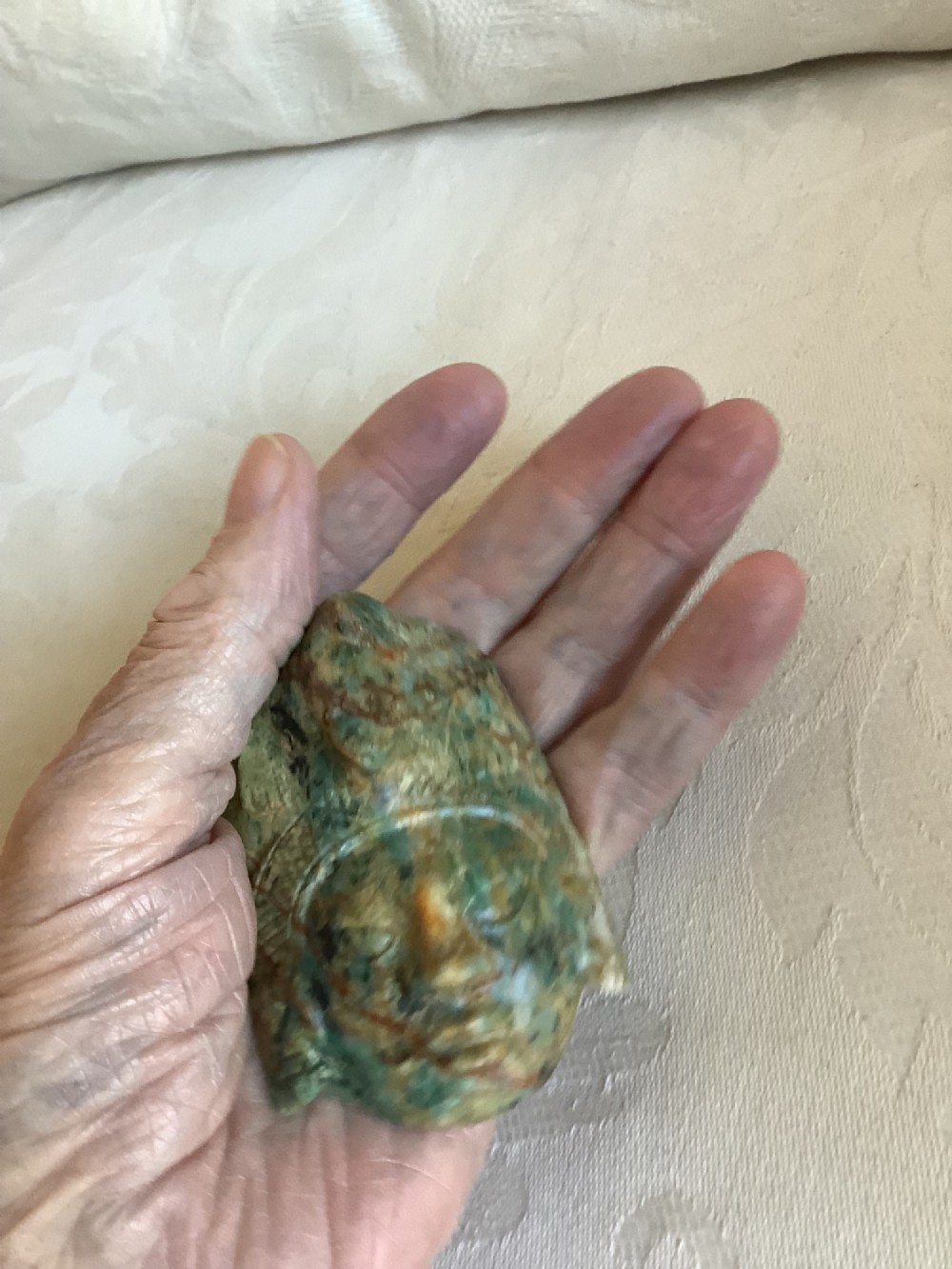 A carved mottled green and brown jade pendant depicting a stylized human face with an ornate carved headdress. View V