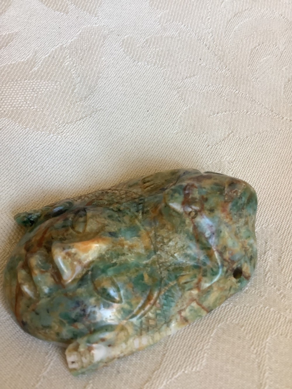 A carved mottled green and brown jade pendant depicting a stylized human face with an ornate carved headdress. View VI