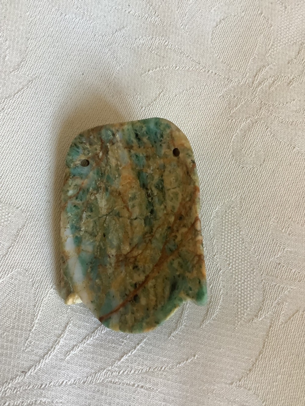 A carved mottled green and brown jade pendant depicting a stylized human face with an ornate carved headdress. View VII