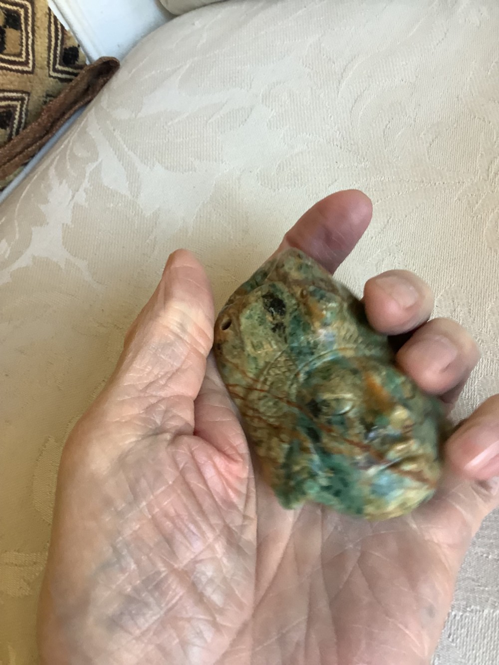 A carved mottled green and brown jade pendant depicting a stylized human face with an ornate carved headdress. View VIII