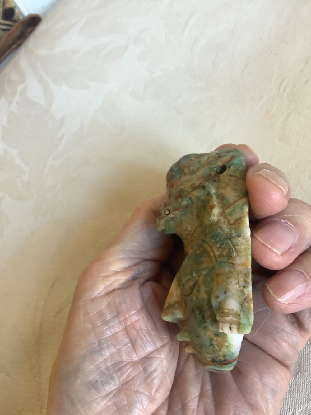 A carved mottled green and brown jade pendant depicting a stylized human face with an ornate carved headdress. View IX