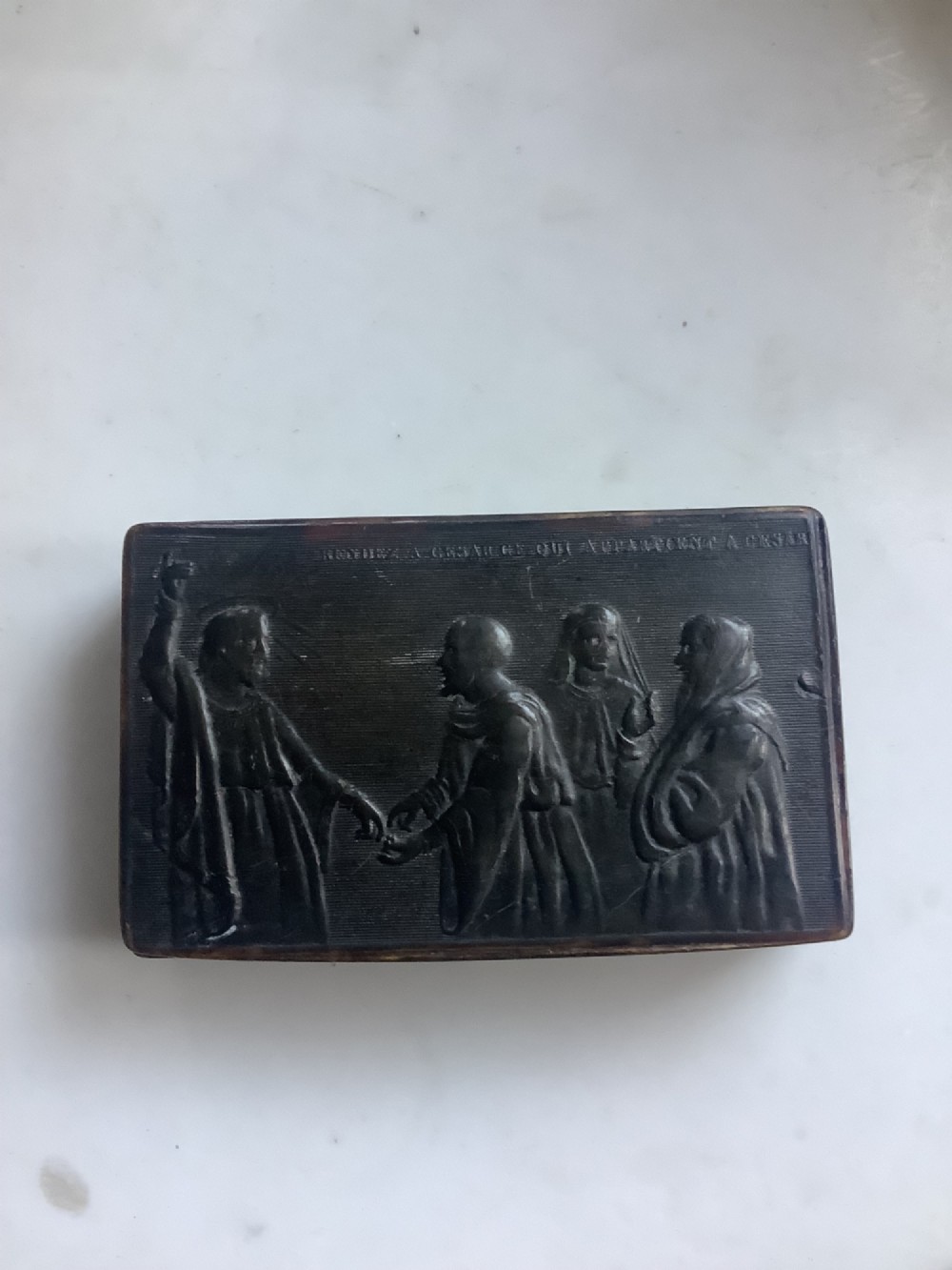 A rectangular dark brown pressed horn snuff box displaying a moulded relief of a biblical scene with figures and a French inscription on the top lid. View II