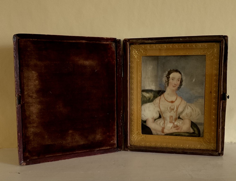 An early Victorian cased portrait miniature depicting a seated lady in a white dress set inside an embossed gilt frame and a velvet-lined burgundy leather folding case.