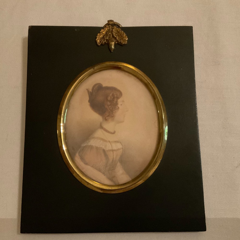A mid-nineteenth century watercolour profile portrait of a young lady set within an oval brass bezel and black rectangular ebonised frame with a top acorn hanger.