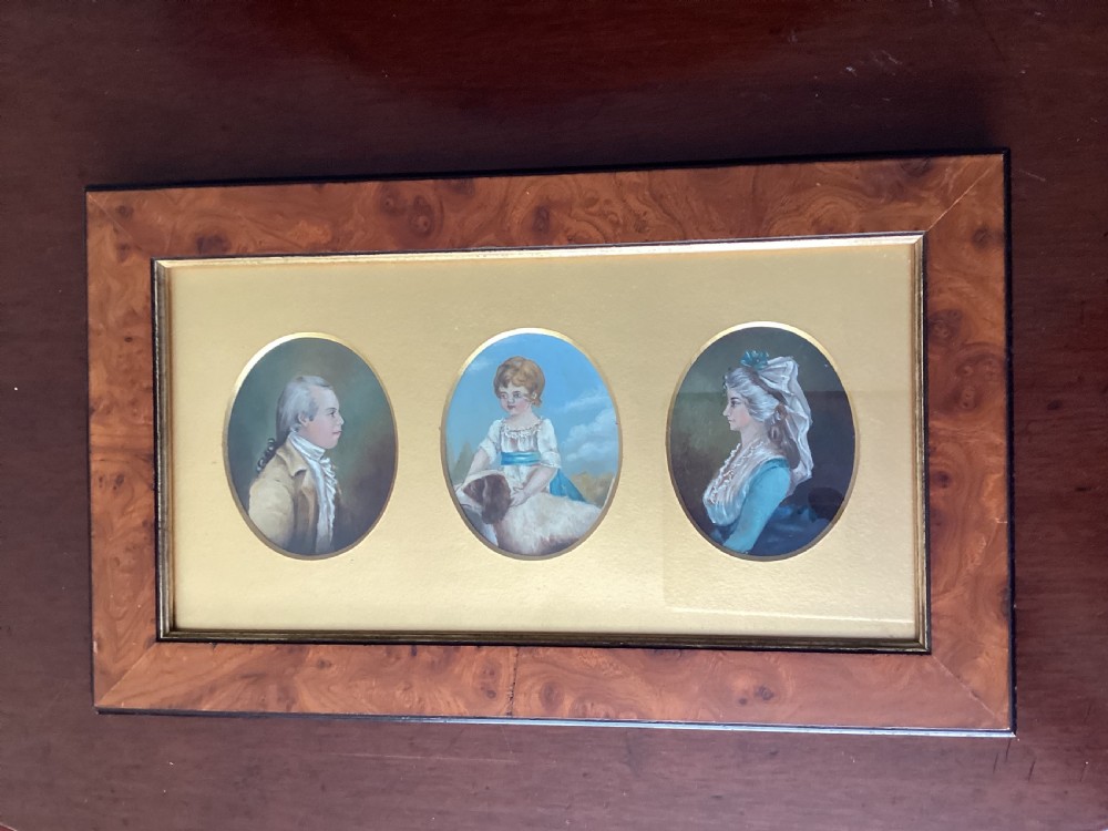 A rectangular burr wood frame enclosing three oval portrait miniatures of a man, a child with a dog, and a woman against a gold mount.