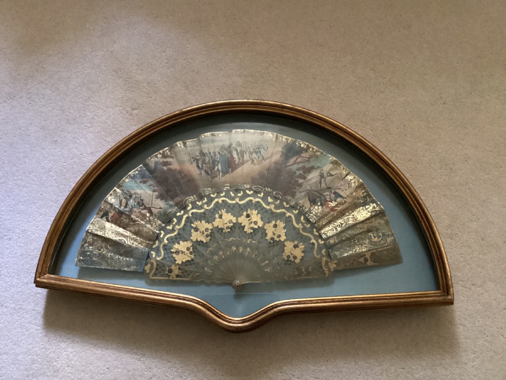 A late nineteenth-century paper folding hand fan decorated with a pastoral scene, presented inside a fan-shaped gilt frame with a blue fabric backing.