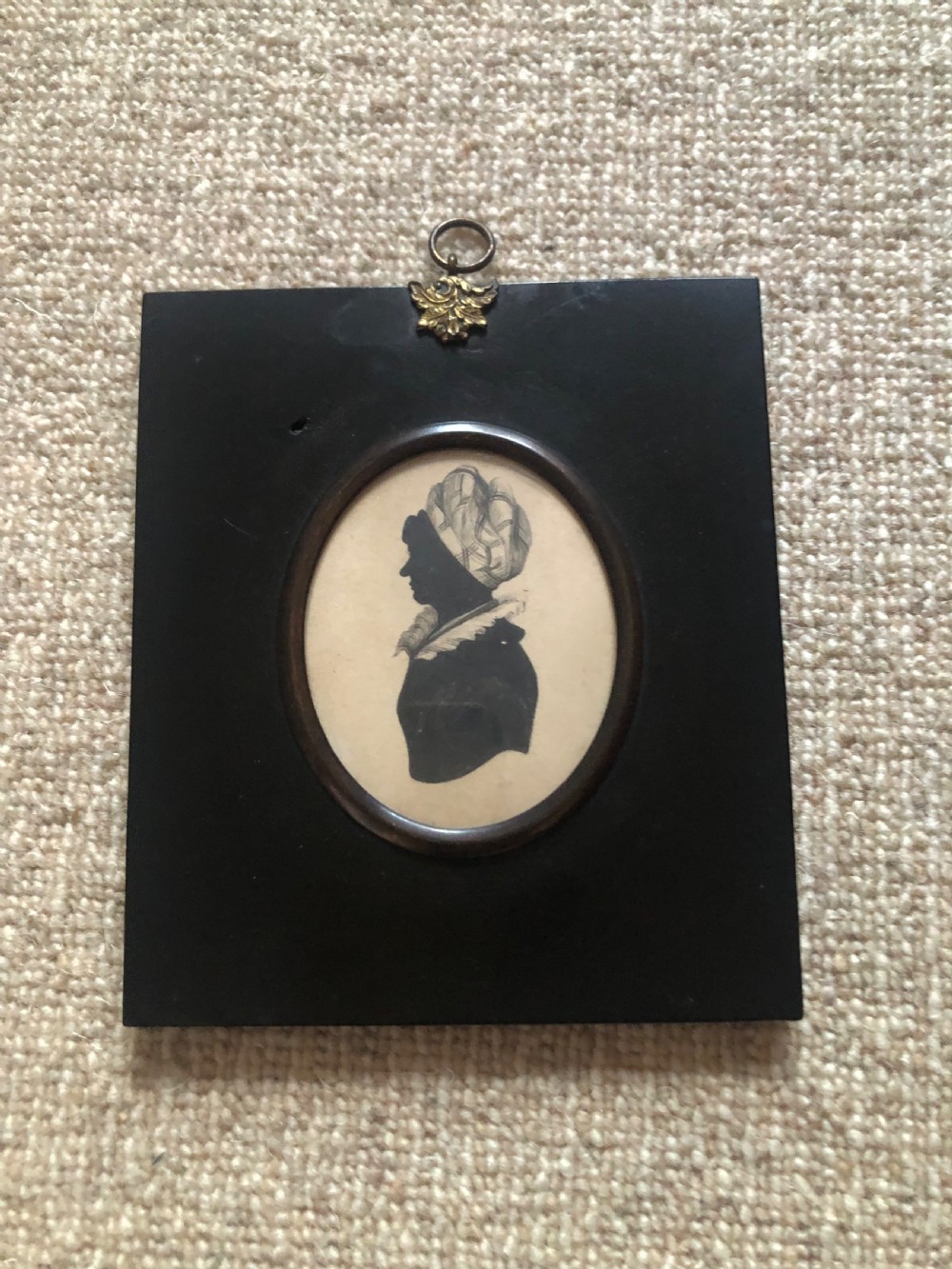 A rectangular black ebonised frame featuring a central oval aperture displaying a black paper silhouette profile of a lady wearing a turban cap, topped with a brass hanging ring.