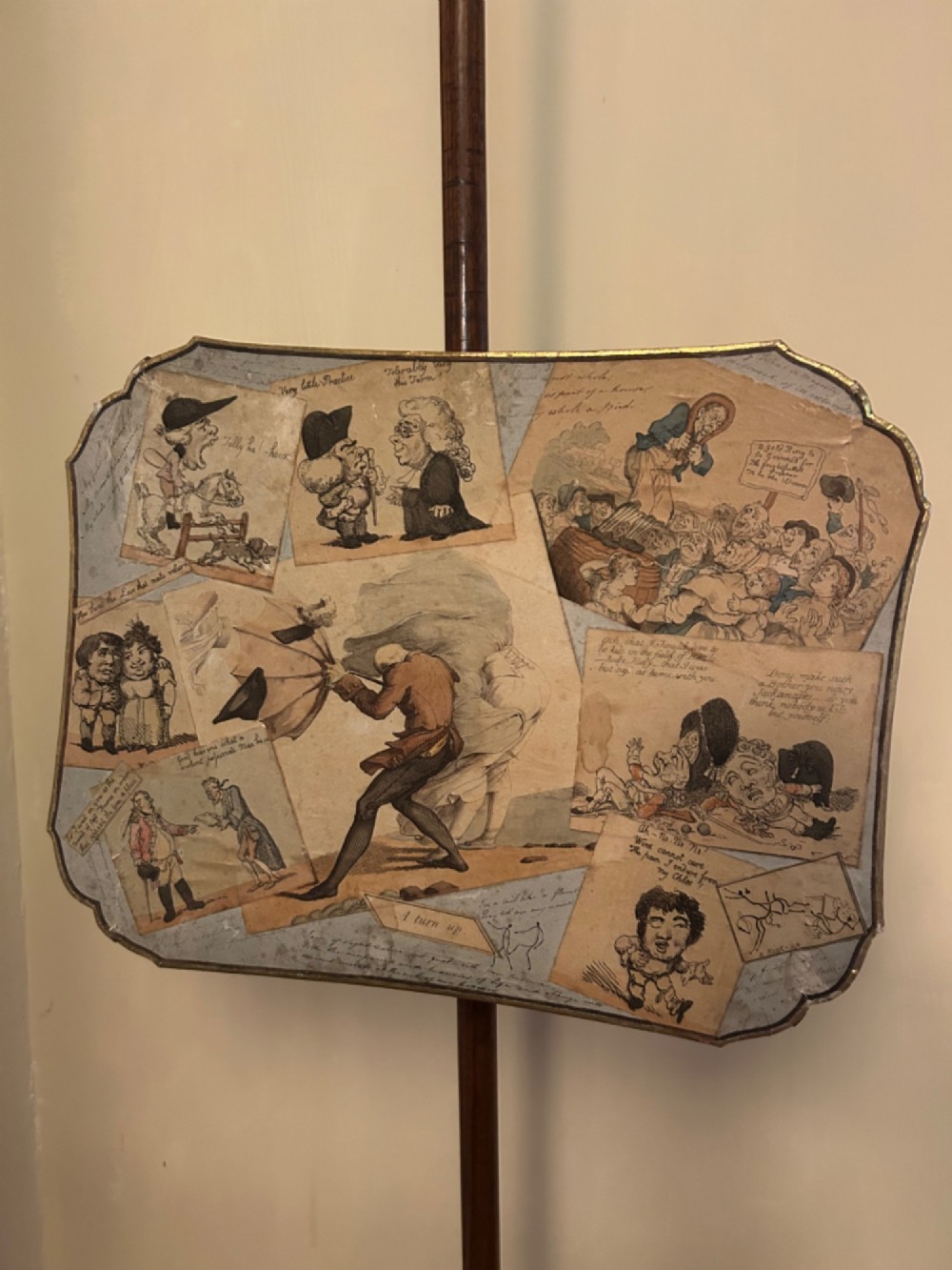 A pair of late George III mahogany pole screens featuring adjustable scalloped panels mounted with printed satirical caricatures on turned wooden stands with tripod bases. View II