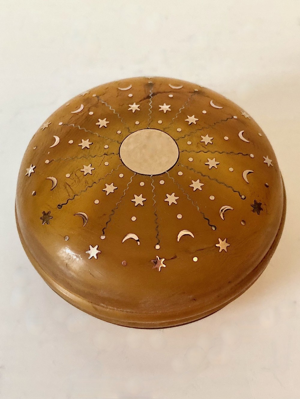 A circular blond tortoiseshell snuff box decorated with celestial inlaid gold stars, crescent moons, and a central metal disc.