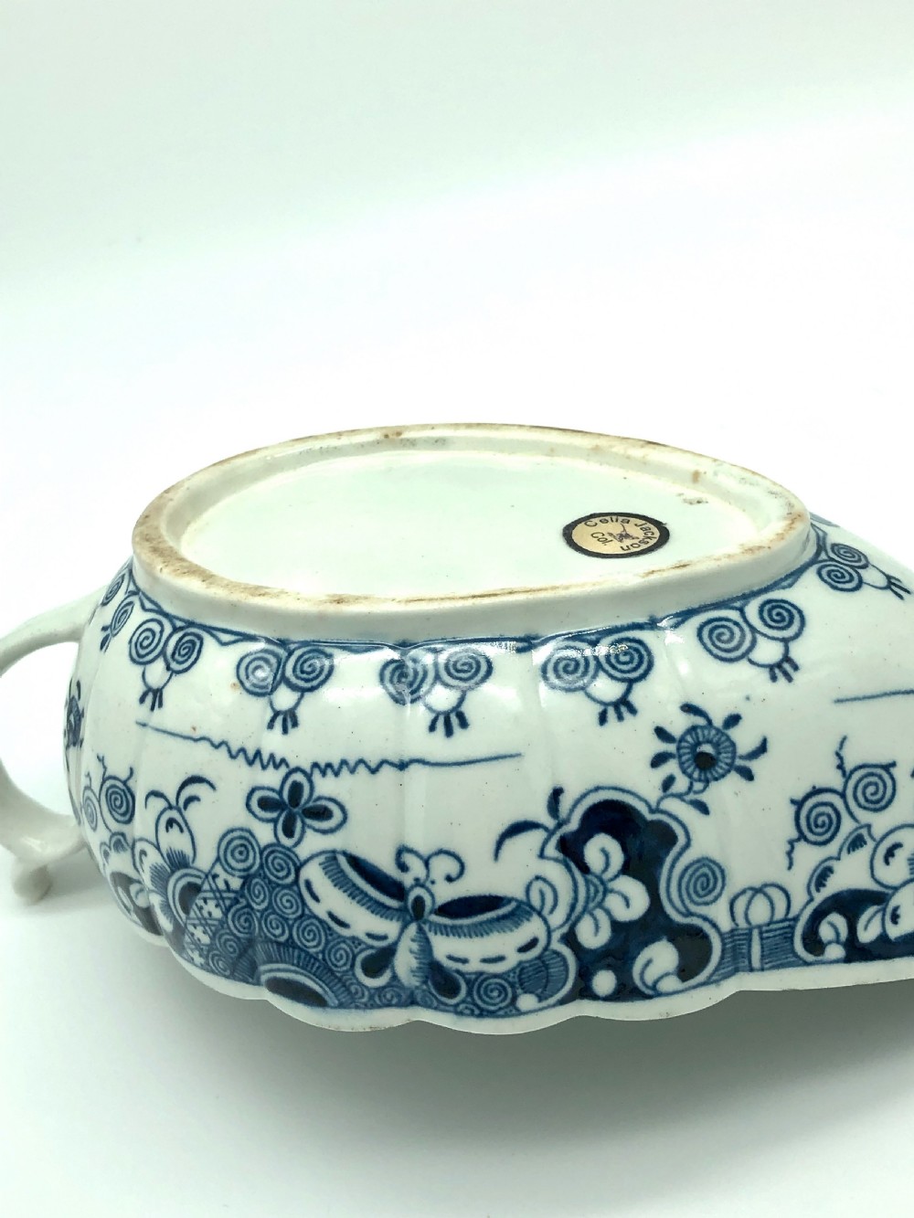 An 18th-century soft-paste porcelain sauceboat decorated with blue and white underglaze motifs, featuring a scalloped rim and a shaped handle. View V