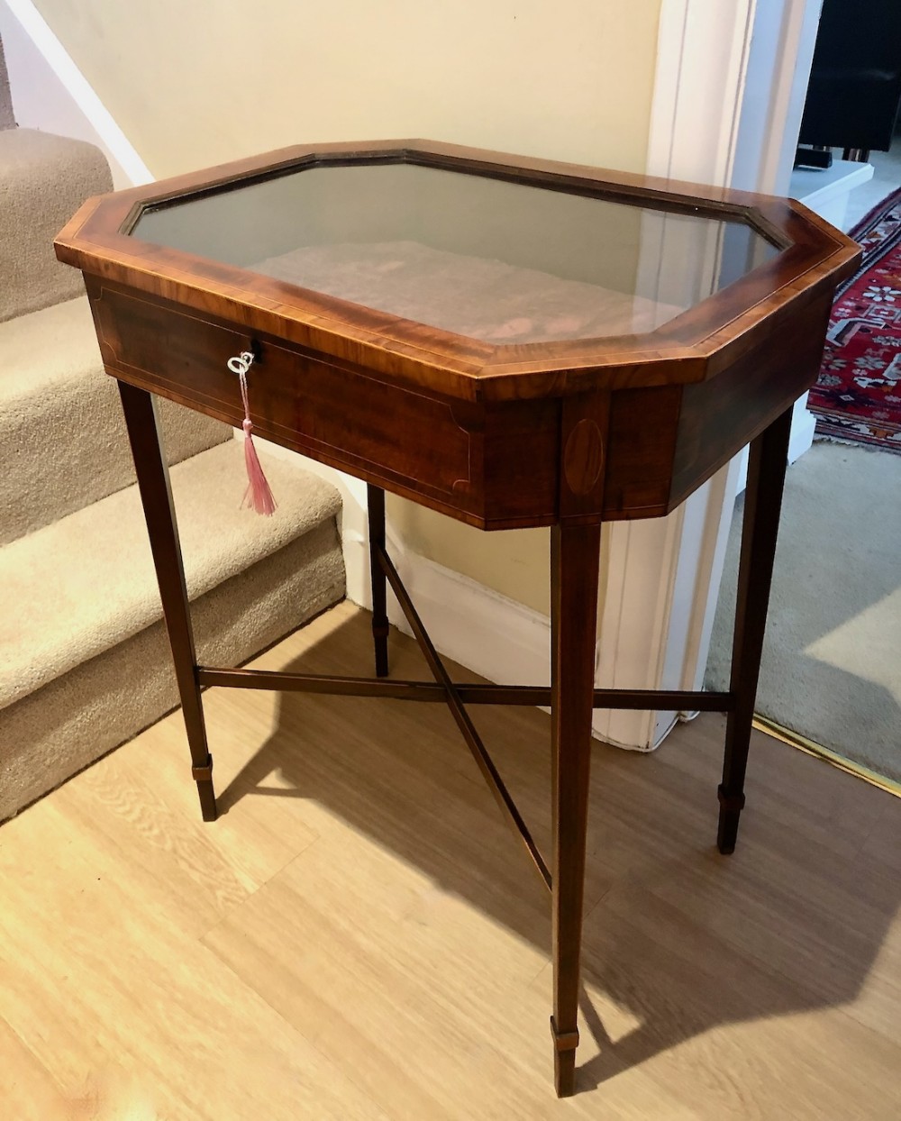 An Edwardian mahogany bijouterie table featuring a canted octagonal glazed top, satinwood stringing inlay, and slender tapered legs with cross stretchers.