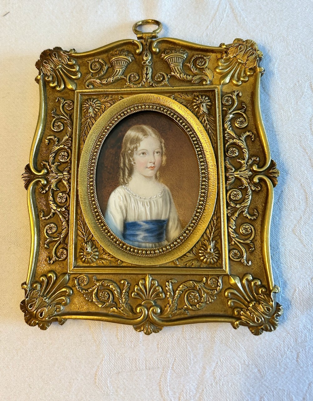 An oval Regency portrait miniature depicting a young girl with fair hair in a white dress and blue sash, encased within an ornate gilt metal rectangular frame. View IV