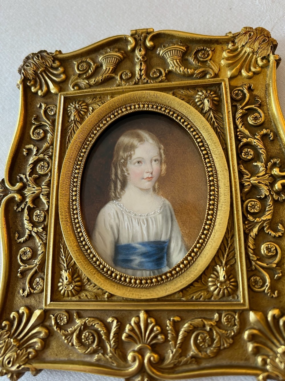 An oval Regency portrait miniature depicting a young girl with fair hair in a white dress and blue sash, encased within an ornate gilt metal rectangular frame. View VI