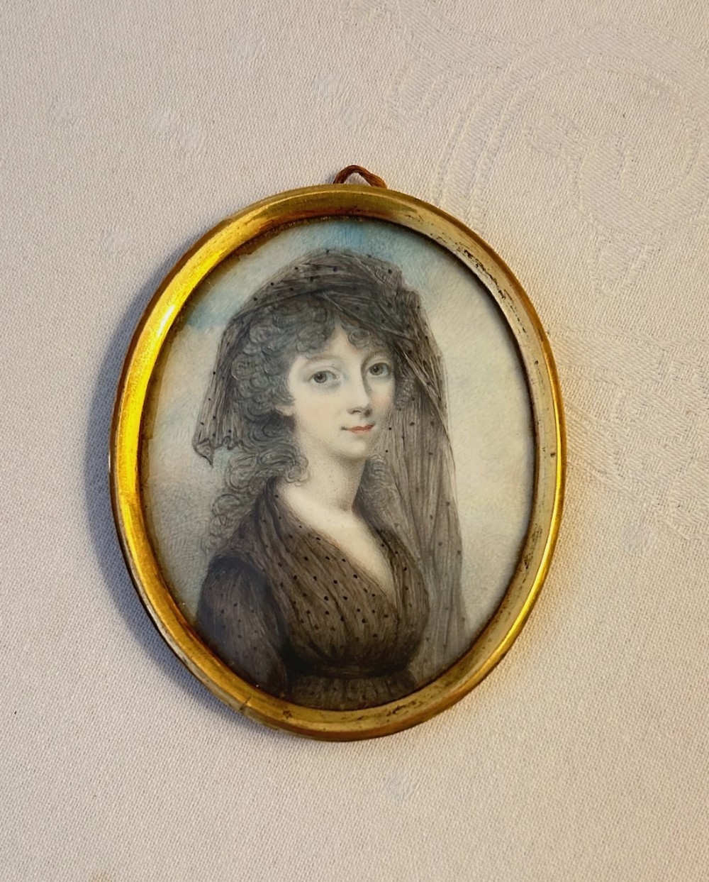 An oval Georgian portrait miniature depicting a woman wearing a laced veil set inside a narrow gilt metal frame with a suspension loop.