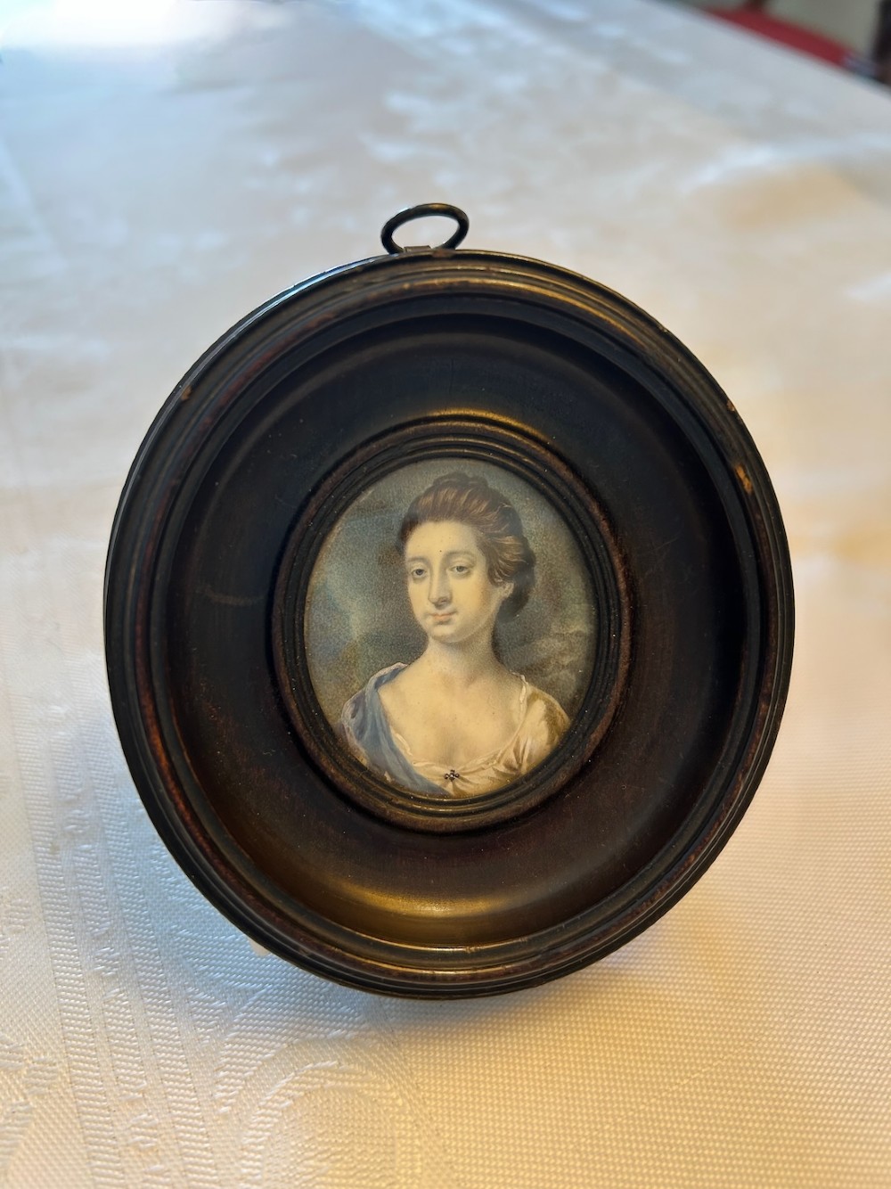 An 18th-century oval portrait miniature depicting a lady in a blue drape, housed within a circular ebonised wood frame with a top metal suspension loop. View IV