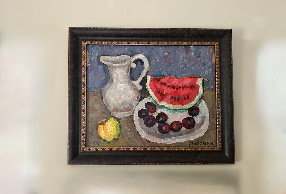 A framed impressionist oil painting depicting a ceramic pitcher, a slice of red watermelon on a plate with dark fruit, and a yellow lemon against a blue background.