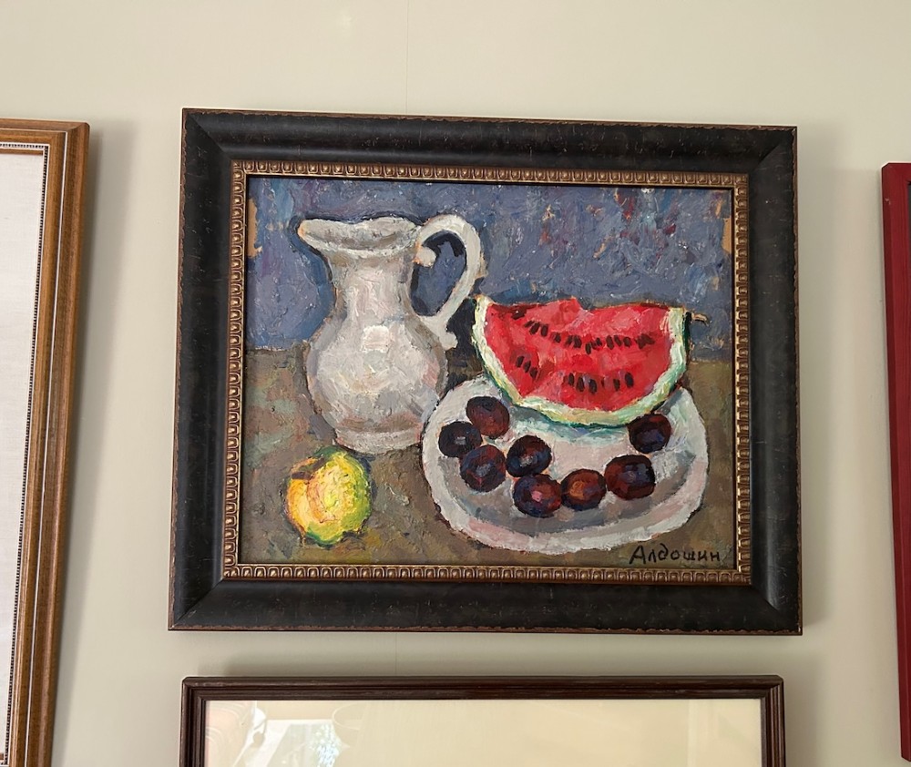 A framed impressionist oil painting depicting a ceramic pitcher, a slice of red watermelon on a plate with dark fruit, and a yellow lemon against a blue background. View II
