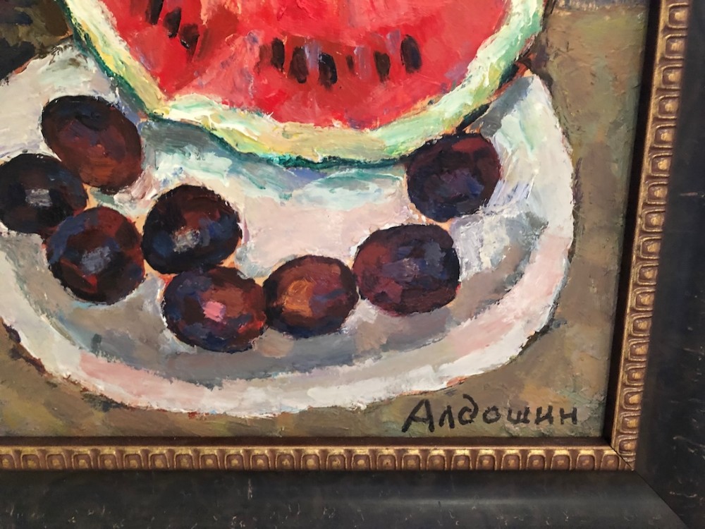 A framed impressionist oil painting depicting a ceramic pitcher, a slice of red watermelon on a plate with dark fruit, and a yellow lemon against a blue background. View III