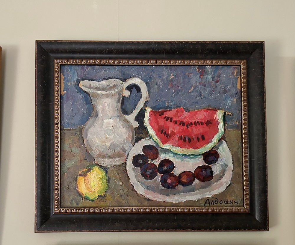 A framed impressionist oil painting depicting a ceramic pitcher, a slice of red watermelon on a plate with dark fruit, and a yellow lemon against a blue background. View IV
