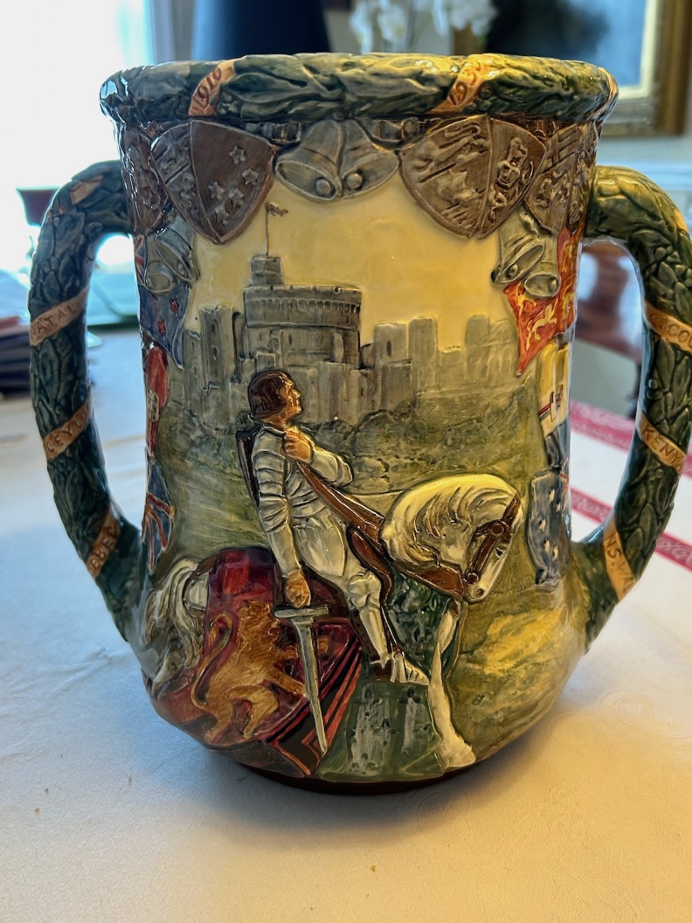 A twin-handled relief-moulded ceramic loving cup displaying profile portraits of King George V and Queen Mary framed by garlands, flags, and heraldic details in warm brown and green glazes. View II