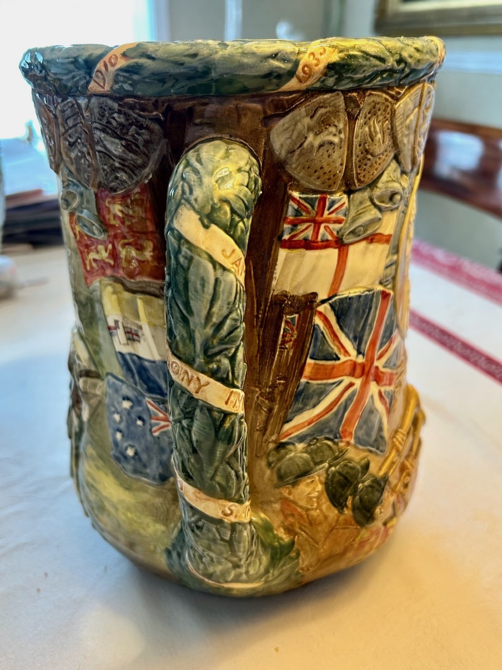 A twin-handled relief-moulded ceramic loving cup displaying profile portraits of King George V and Queen Mary framed by garlands, flags, and heraldic details in warm brown and green glazes. View III