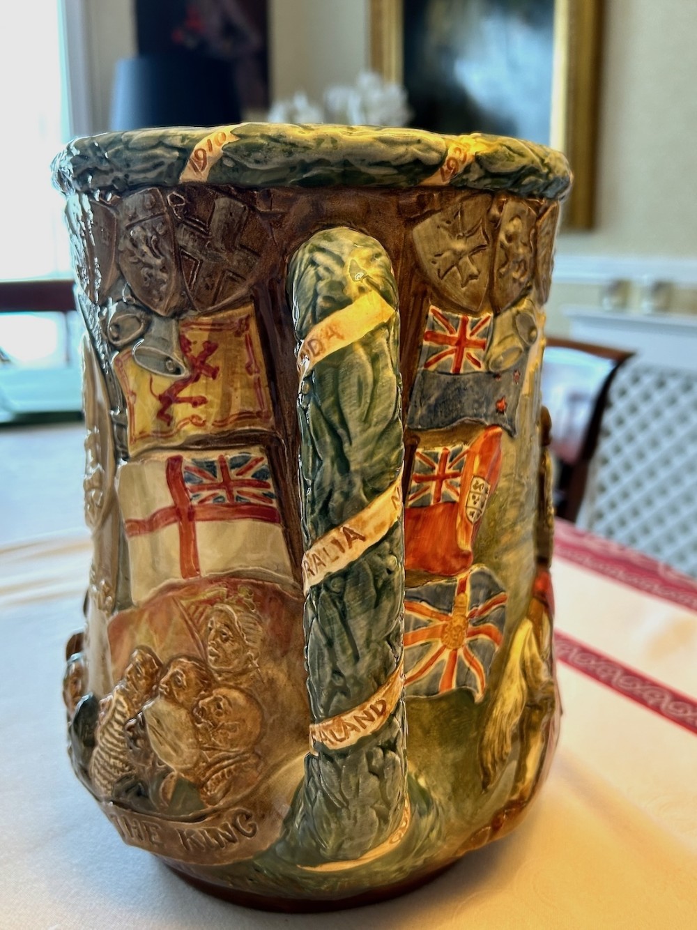 A twin-handled relief-moulded ceramic loving cup displaying profile portraits of King George V and Queen Mary framed by garlands, flags, and heraldic details in warm brown and green glazes. View IV