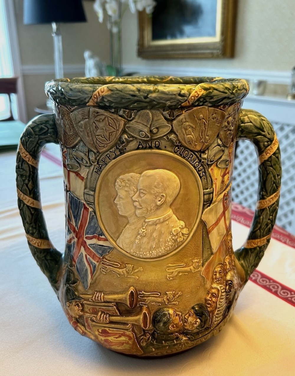 A twin-handled relief-moulded ceramic loving cup displaying profile portraits of King George V and Queen Mary framed by garlands, flags, and heraldic details in warm brown and green glazes. View V