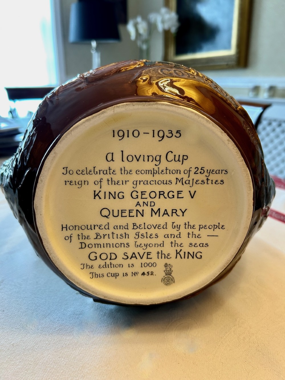 A twin-handled relief-moulded ceramic loving cup displaying profile portraits of King George V and Queen Mary framed by garlands, flags, and heraldic details in warm brown and green glazes. View VI