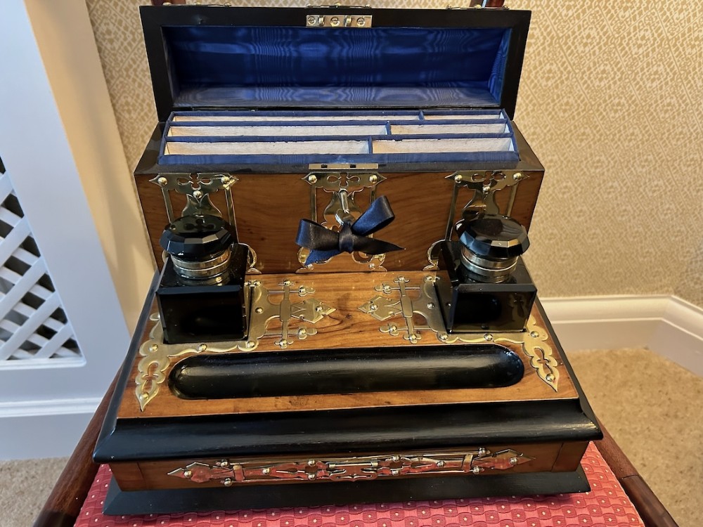 An open Victorian figured walnut desk stand featuring decorative brass strapwork, two black inkwells, a pen trough, and an upper stationery compartment lined in blue fabric. View V