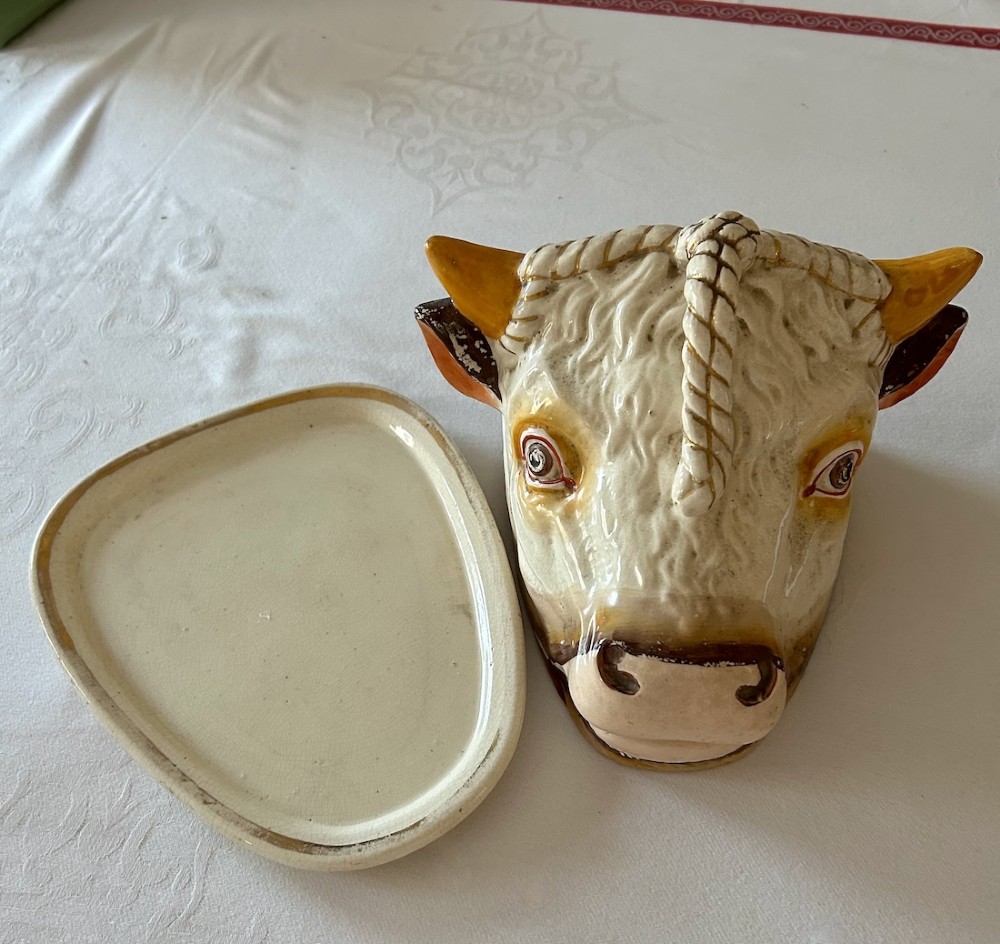 An antique 19th-century glazed cream ceramic cheese dish shaped like a cow head with yellow horns resting on an oval base. View II