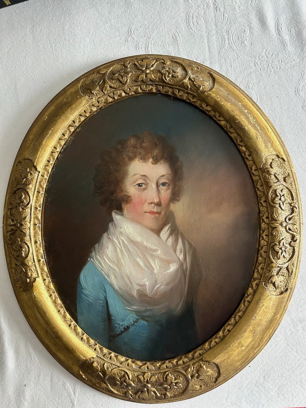 An oval Regency-era oil painting depicting a lady in a blue dress and white scarf, housed in an ornate giltwood frame.