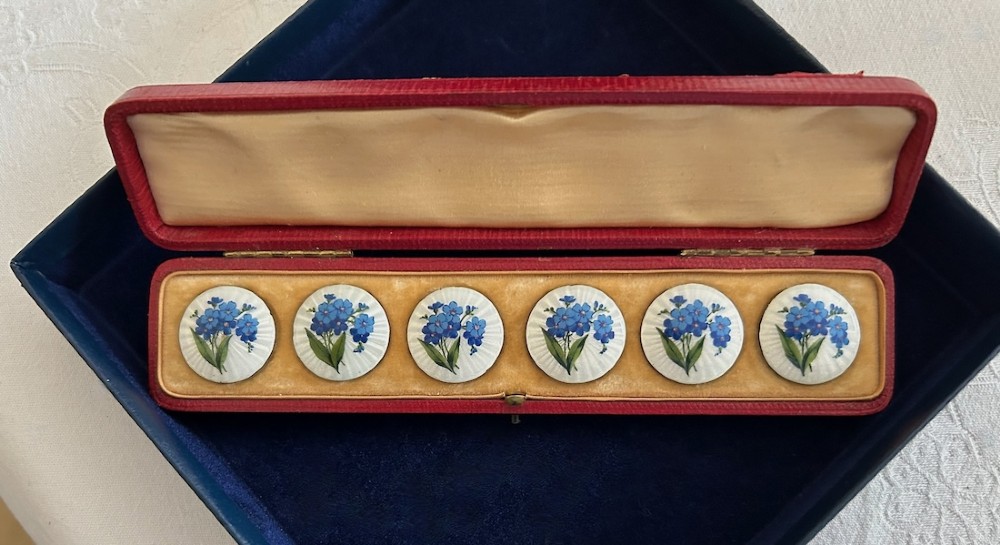 A set of six circular Edwardian silver buttons decorated with blue forget-me-not flowers on white guilloche enamel, displayed inside a fitted red presentation case.