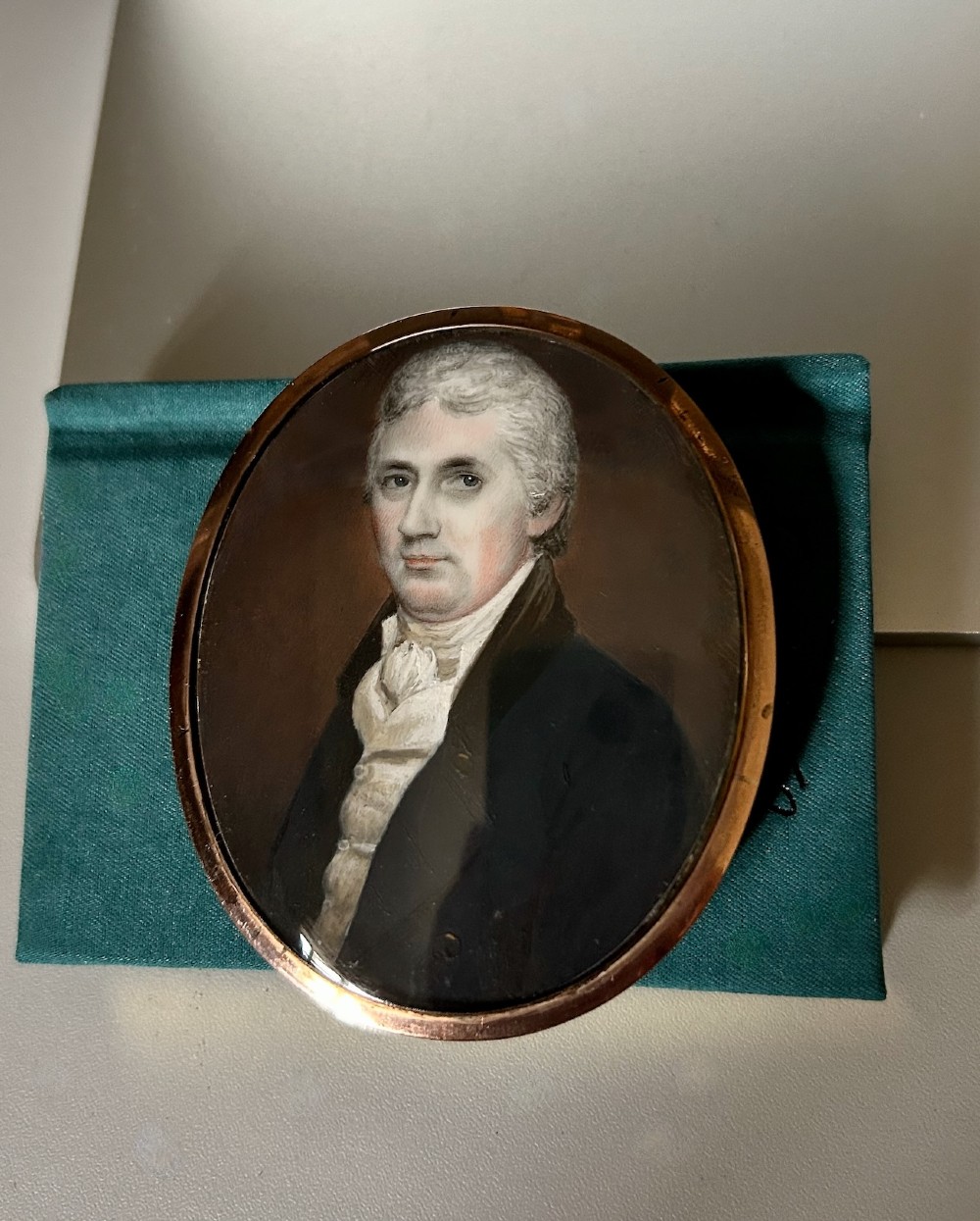 An oval Georgian portrait miniature depicting a gentleman with grey hair wearing a dark coat and white cravat, housed within a gold-plated frame.
