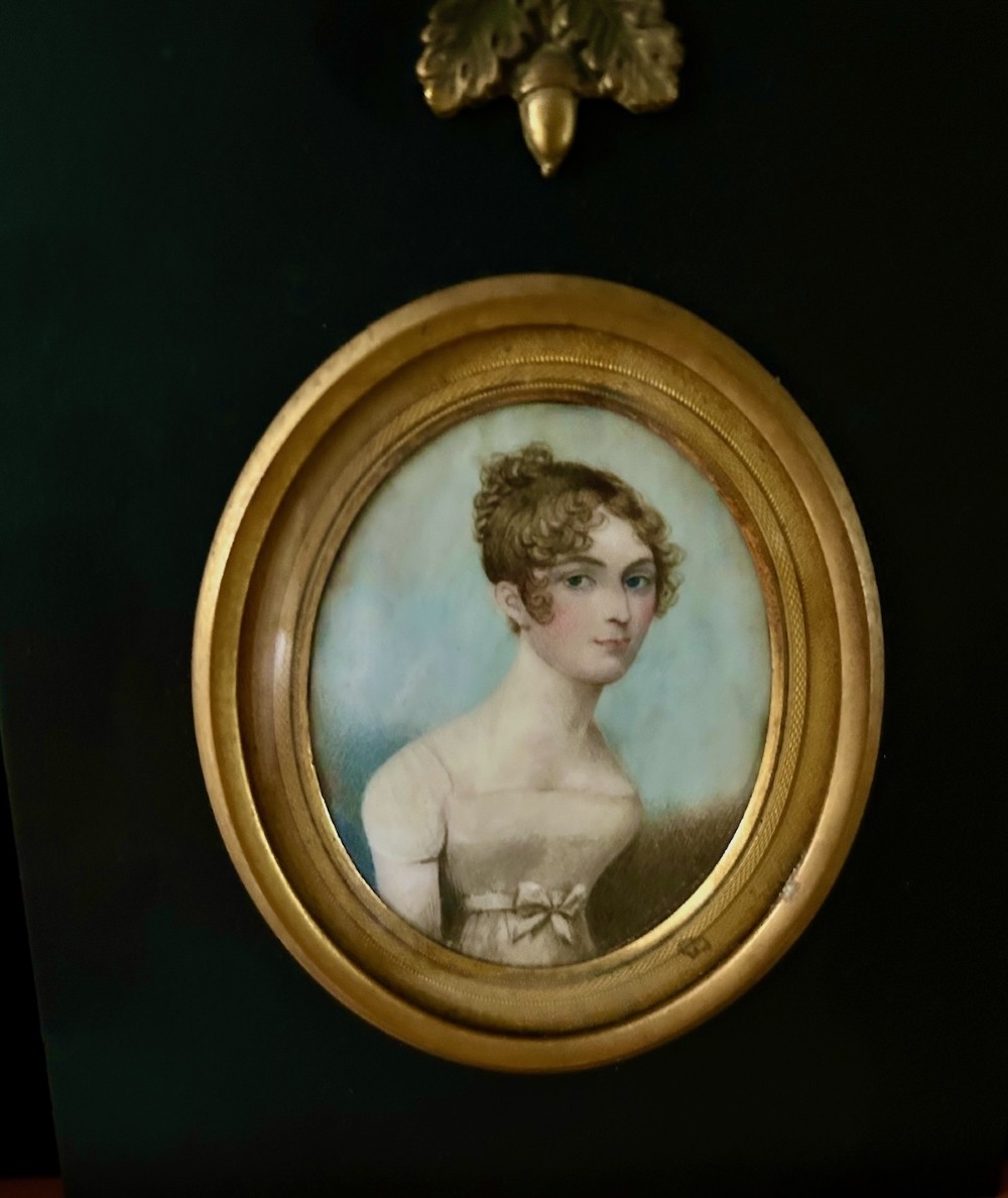 An oval early nineteenth-century watercolour portrait miniature depicting a young lady with curled brown hair, framed in a gilt bezel set within a black ebonised mount featuring a brass acorn top ornament.