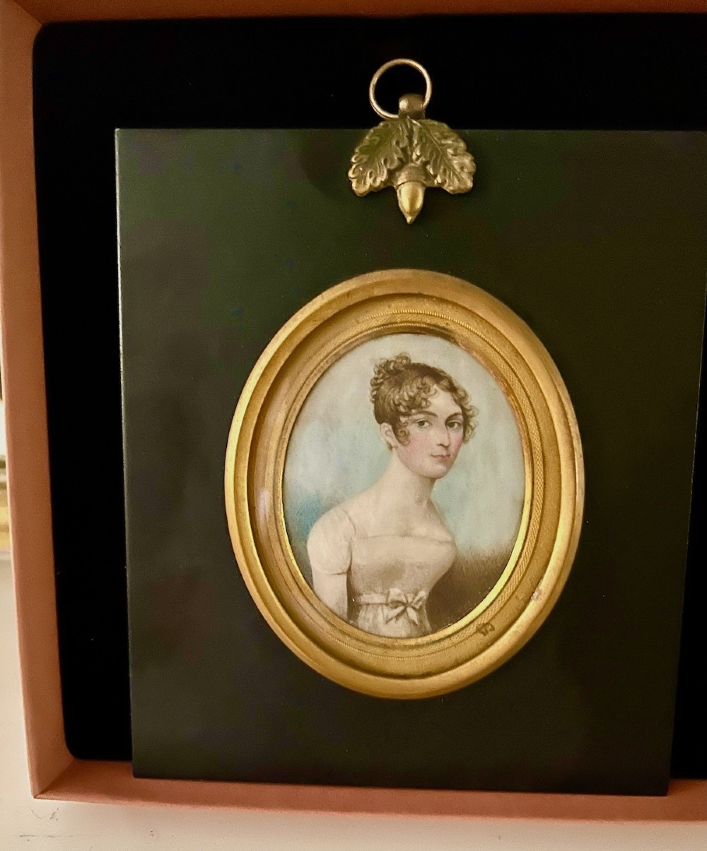 An oval early nineteenth-century watercolour portrait miniature depicting a young lady with curled brown hair, framed in a gilt bezel set within a black ebonised mount featuring a brass acorn top ornament. View II