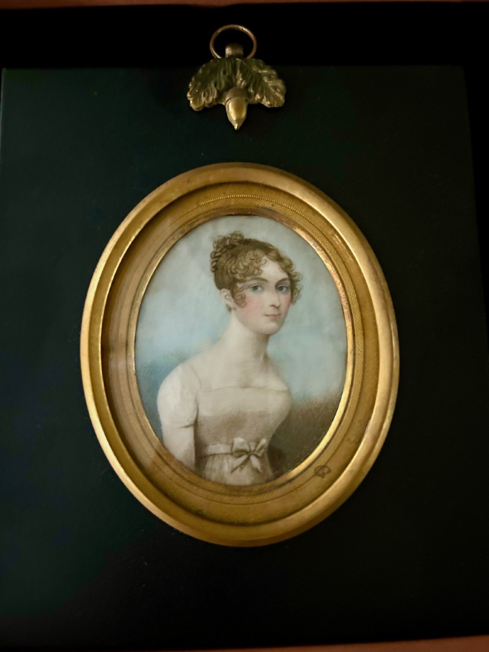 An oval early nineteenth-century watercolour portrait miniature depicting a young lady with curled brown hair, framed in a gilt bezel set within a black ebonised mount featuring a brass acorn top ornament. View III