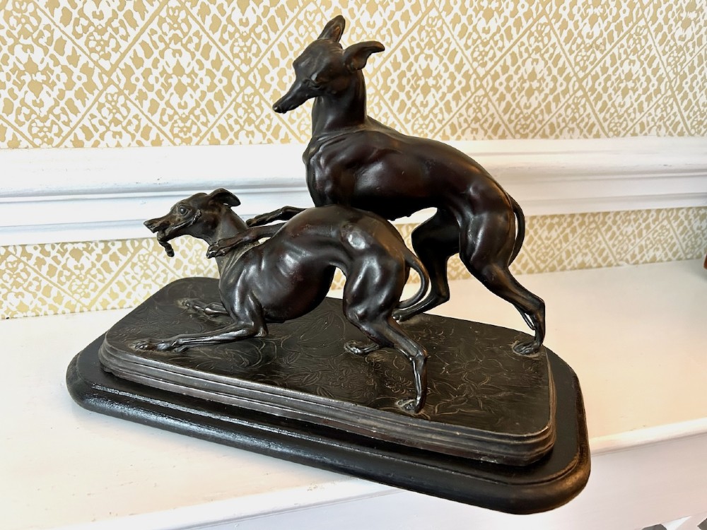 A dark brown patinated spelter sculpture depicting two whippets on a detailed relief base mounted upon a moulded wooden plinth.