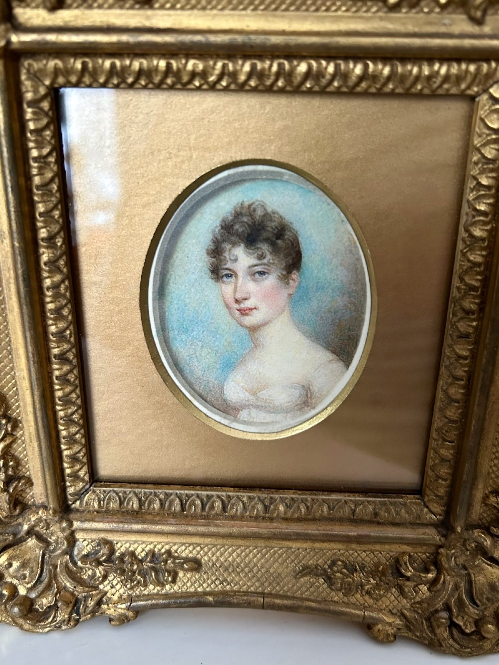An early nineteenth-century oval watercolour portrait miniature of a lady in a white gown set within a rectangular carved giltwood frame. View IV