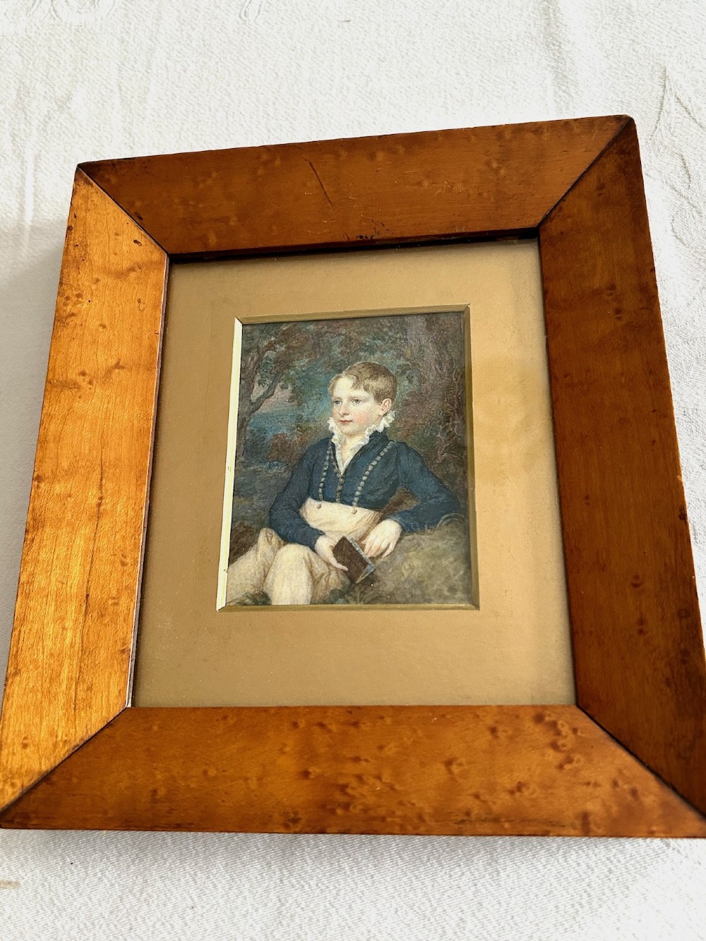A Victorian watercolour portrait depicting a young boy holding a book seated outdoors, displayed inside a wide birdseye maple wooden frame. View II