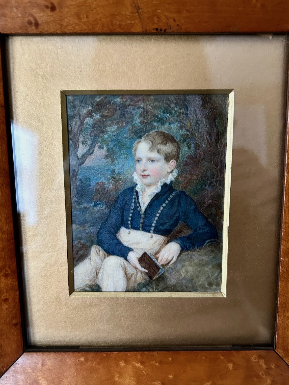 A Victorian watercolour portrait depicting a young boy holding a book seated outdoors, displayed inside a wide birdseye maple wooden frame. View III
