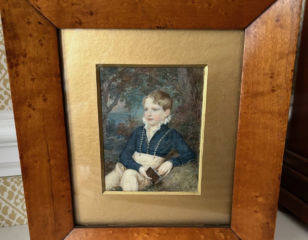 A Victorian watercolour portrait depicting a young boy holding a book seated outdoors, displayed inside a wide birdseye maple wooden frame. View V