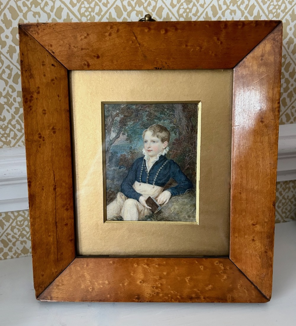 A Victorian watercolour portrait depicting a young boy holding a book seated outdoors, displayed inside a wide birdseye maple wooden frame. View VI